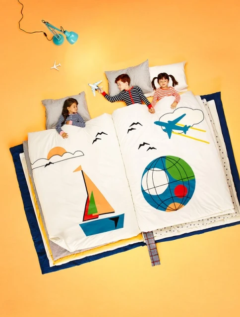 Overhead shot of three children lying on a large book-shaped bed with airplane and sailboat illustrations, creating a whimsical travel theme.
