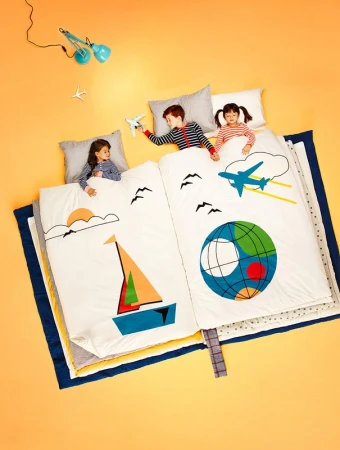 Overhead shot of three children lying on a large book-shaped bed with airplane and sailboat illustrations, creating a whimsical travel theme.