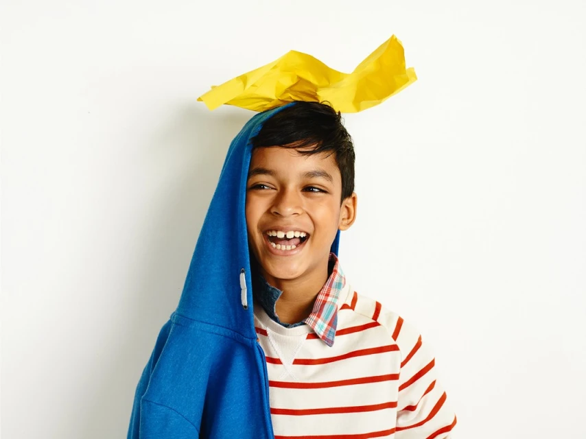 Boy with blue hoodie and yellow paper on his head smiles against a white background in a bright studio lighting setting.