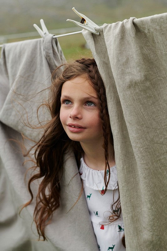 A young girl with braided brown hair peers from behind neutral-colored linen sheets on a clothesline outdoors in soft, diffused light.