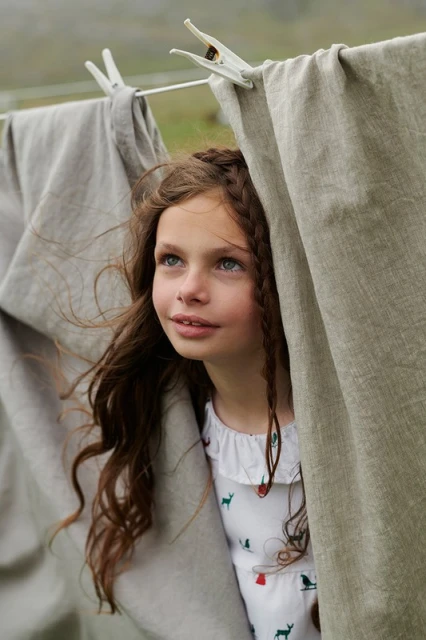 A young girl with braided brown hair peers from behind neutral-colored linen sheets on a clothesline outdoors in soft, diffused light.