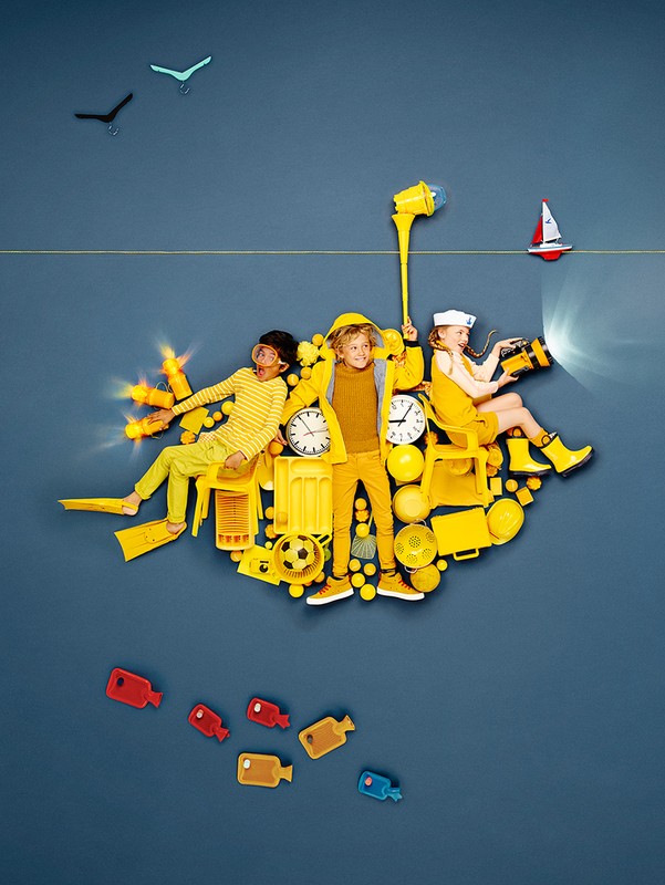 Three playful children on a yellow object island against a dark blue background with bird figures on the wall and a boat figure.