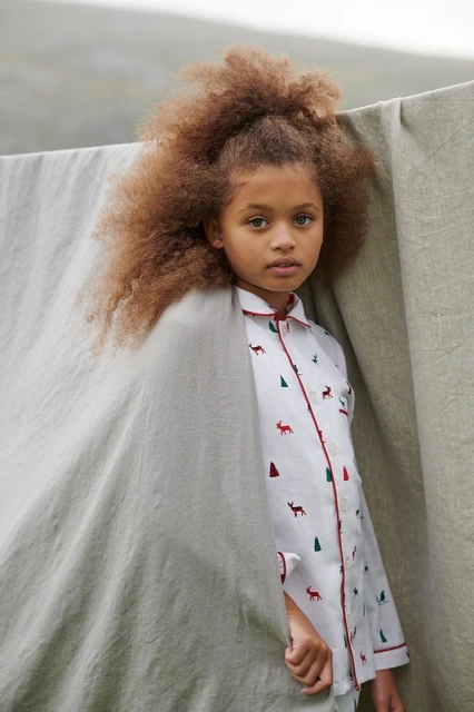 Young child with afro hairstyle, wearing a patterned pajama top, poses near a grey linen cloth outdoors, in a medium portrait shot.