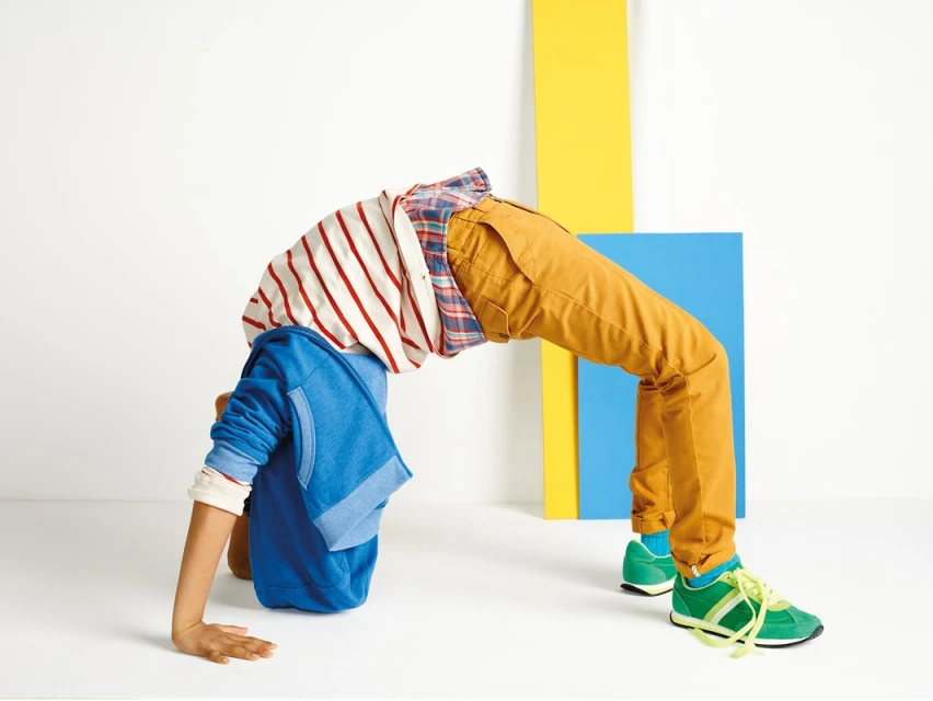 Young boy wearing a blue hooded sweatshirt, striped shirt, and orange pants, doing a backbend in a white studio setting.
