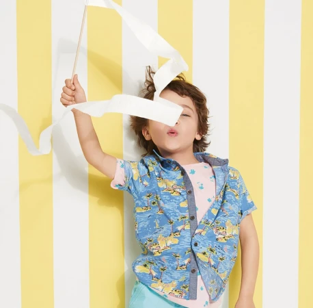A young child poses with a white ribbon against a yellow and white striped background, wearing a patterned shirt and mint-green shorts.