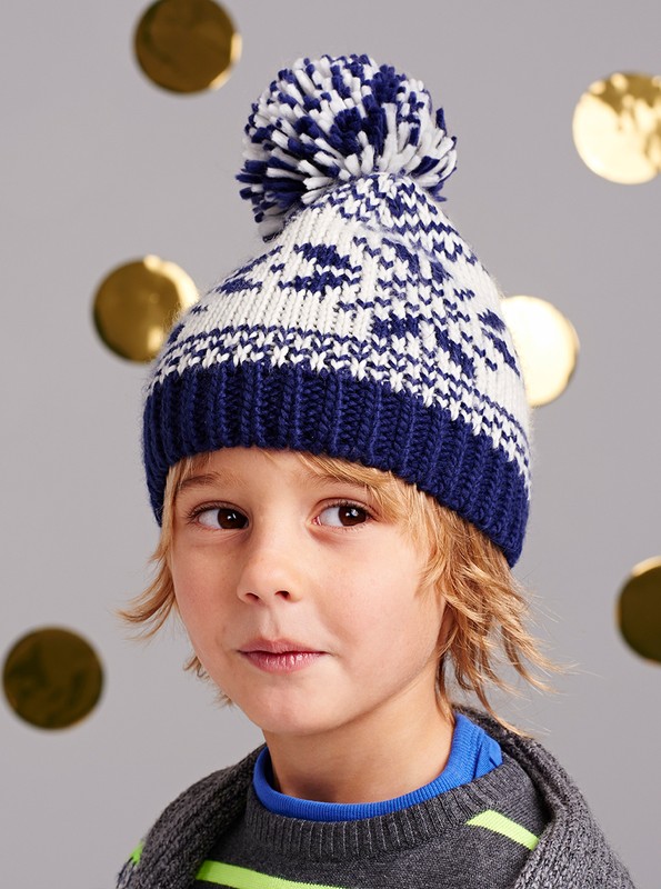 Young boy with blonde hair wearing a blue and white winter hat with a pompom, dressed in a grey sweater against a grey background.