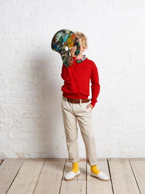 Young boy in red sweater holds a paint palette in front of his face indoors, wearing tan pants, yellow socks, and white shoes.