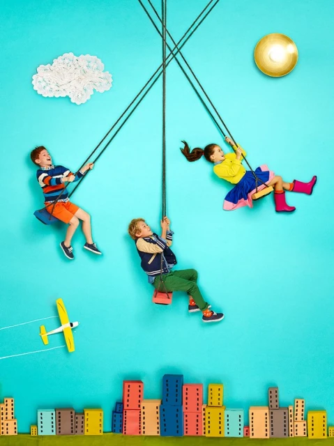 Three children on colorful swings against a light blue background with building blocks and a white cloud art installation.