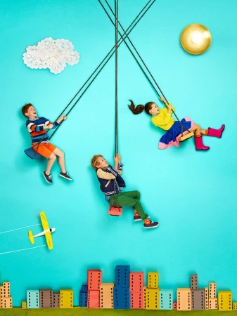 Three children on colorful swings against a light blue background with building blocks and a white cloud art installation.