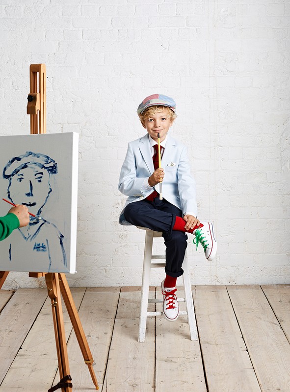 Blond boy in cap sits with a paintbrush, an unfinished portrait on a wooden easel with a green hand using a brush.