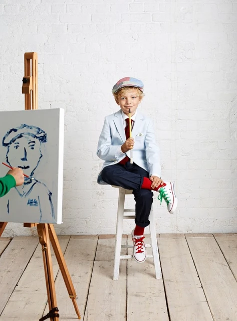 Blond boy in cap sits with a paintbrush, an unfinished portrait on a wooden easel with a green hand using a brush.