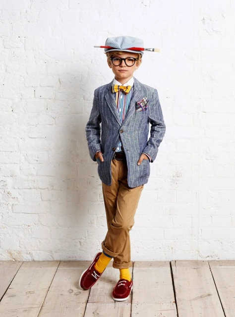 Full shot of a child dressed in a blazer, striped shirt, bow tie, glasses, hat, and trousers, standing against a white brick wall.