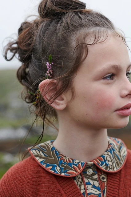 Young girl with flowers tucked behind her ear in an outdoor setting, wearing a floral-patterned shirt and orange cardigan.