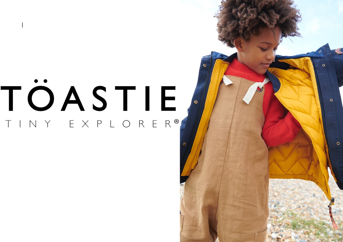 Child wearing Toastie Tiny Explorer outfit, including tan overalls and a blue jacket with a yellow quilted lining, outdoors.