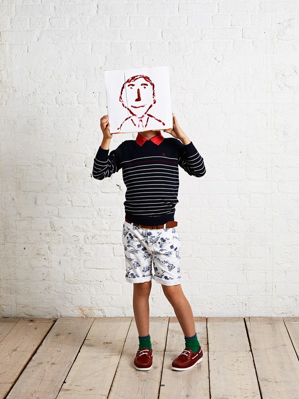 A child model wearing patterned shorts and a striped sweater holds up a drawing of a smiling face against a white brick wall indoors.