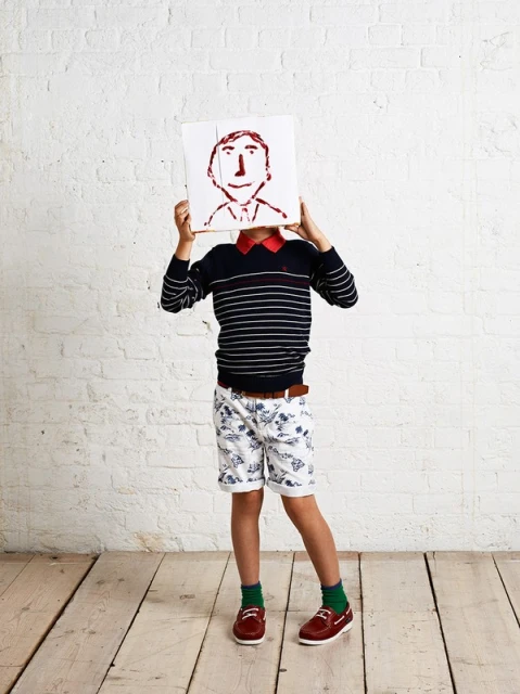 A child model wearing patterned shorts and a striped sweater holds up a drawing of a smiling face against a white brick wall indoors.