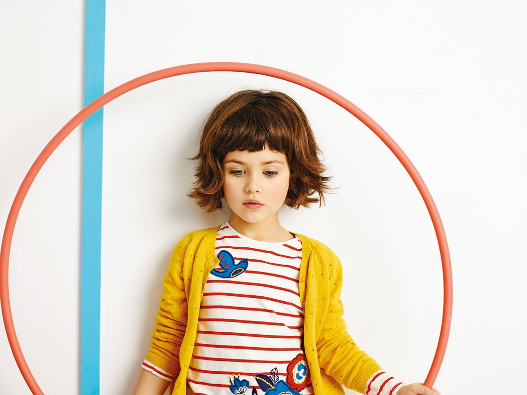 A young girl with short brown hair poses indoors in a striped shirt and yellow cardigan, holding a hula hoop against a white backdrop.