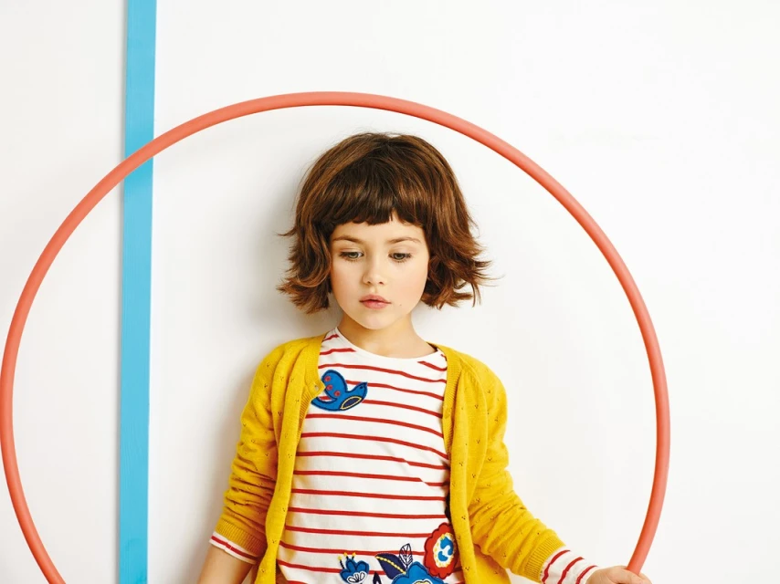 A young girl with short brown hair poses indoors in a striped shirt and yellow cardigan, holding a hula hoop against a white backdrop.