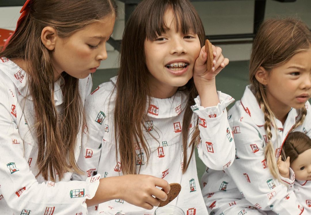 Three young girls in matching pajamas sit together, one holding a cookie. A doll and a glass of milk are visible.