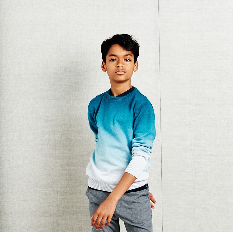 Teenage boy stands in a blue and white ombré sweater and gray pants in front of a neutral-colored wall in a studio shot.
