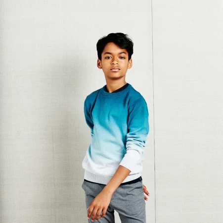 Teenage boy stands in a blue and white ombré sweater and gray pants in front of a neutral-colored wall in a studio shot.