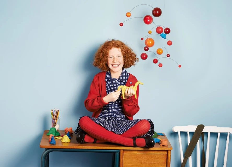 A girl with red hair sits cross-legged on a wooden desk, playfully holding a yellow toy dinosaur in a light blue room.