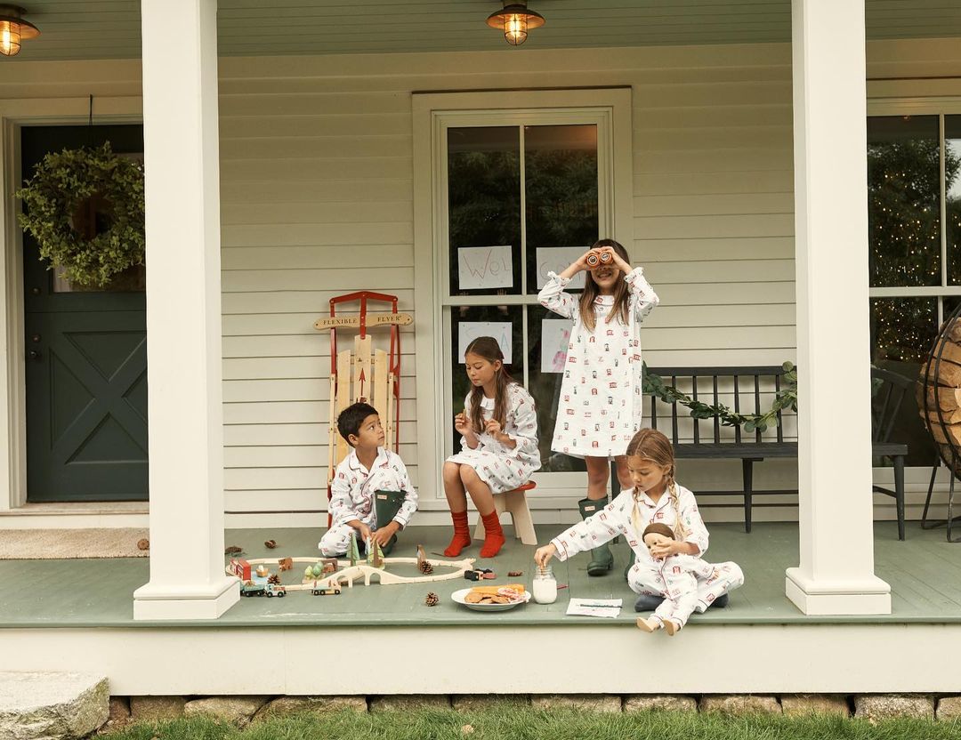 Four children in matching pyjamas play on a porch with toys, holiday decorations, and a red sled against a light-colored house.