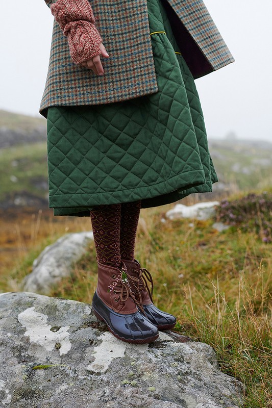 A person stands on a grey rock wearing brown duck boots, patterned tights, a green quilted skirt, and a plaid jacket in a misty, rural setting.