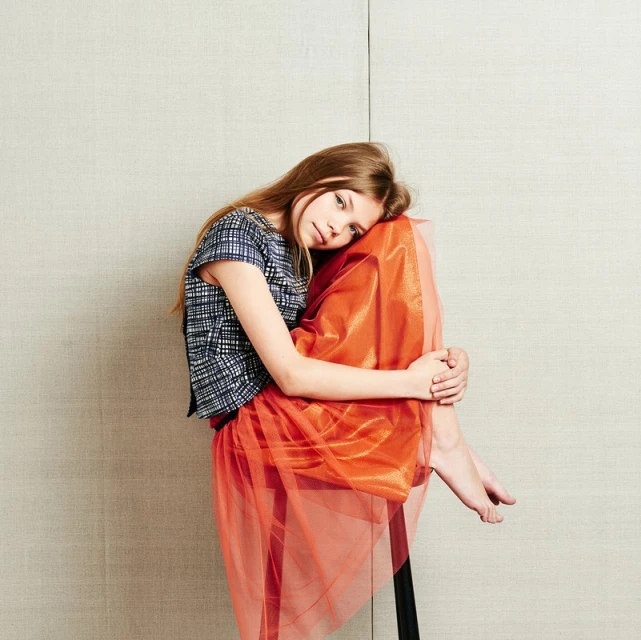 Young girl with long blonde hair sits and hugs her knees in a photo studio wearing a patterned short-sleeve shirt and orange skirt.