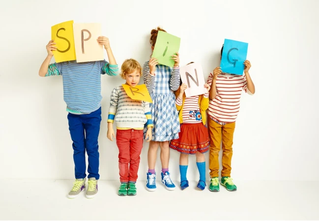 Five children stand shoulder-to-shoulder in front of a white wall holding colorful paper letters that spell out the word SPRING.