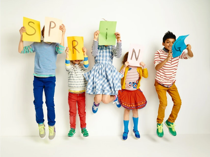 Five children jumping in the air holding up colorful letters that spell SPRING, wearing colorful casual outfits against a white background.
