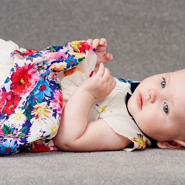 Infant lying down wearing a floral dress in a studio setting. The baby is looking directly at the camera with a neutral expression.