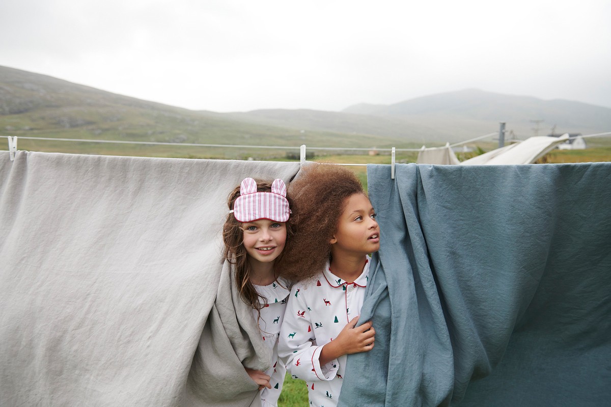 Two young girls in pajamas standing behind draped laundry outdoors with a mountain backdrop, wearing sleep masks and smiling.