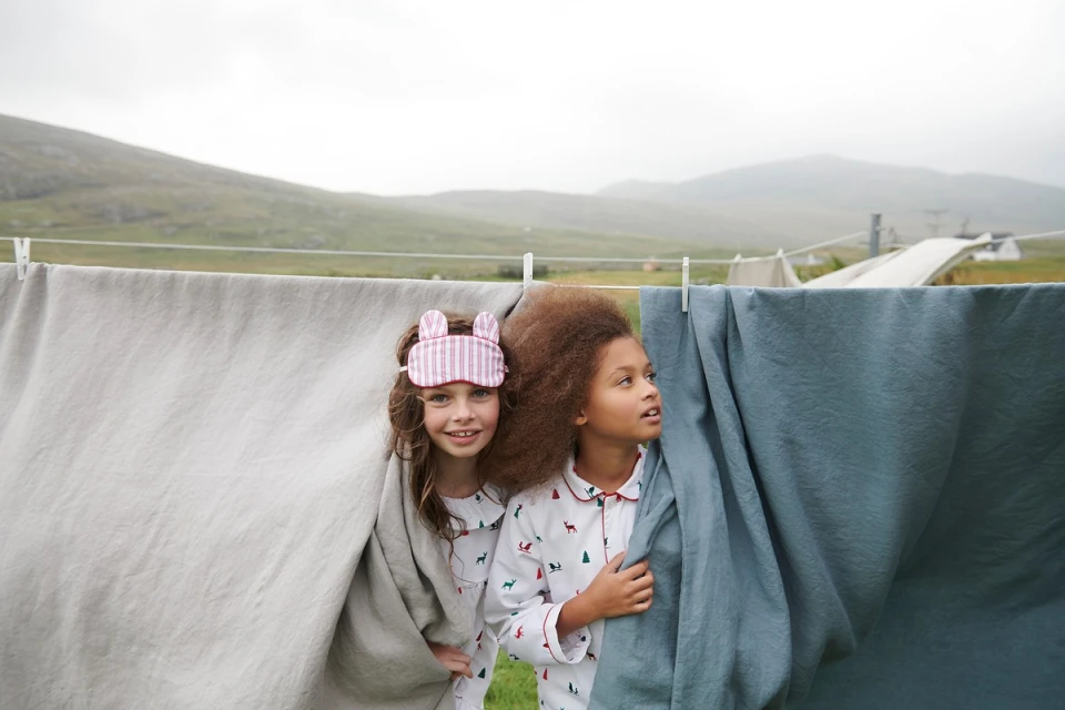Two young girls in pajamas standing behind draped laundry outdoors with a mountain backdrop, wearing sleep masks and smiling.