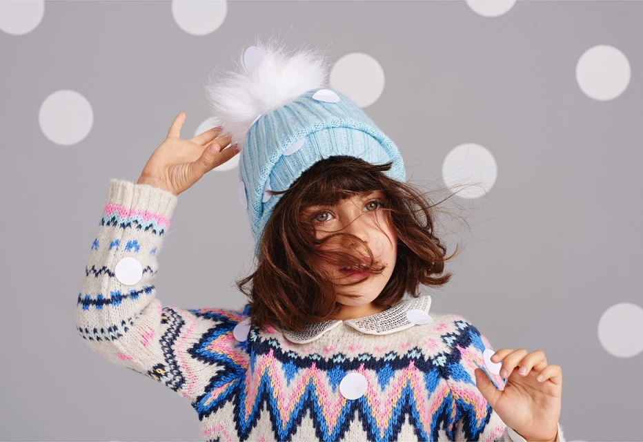 Adorable young girl in a colorful sweater and blue pom-pom hat poses in a studio with polka dot background, looking slightly upward.