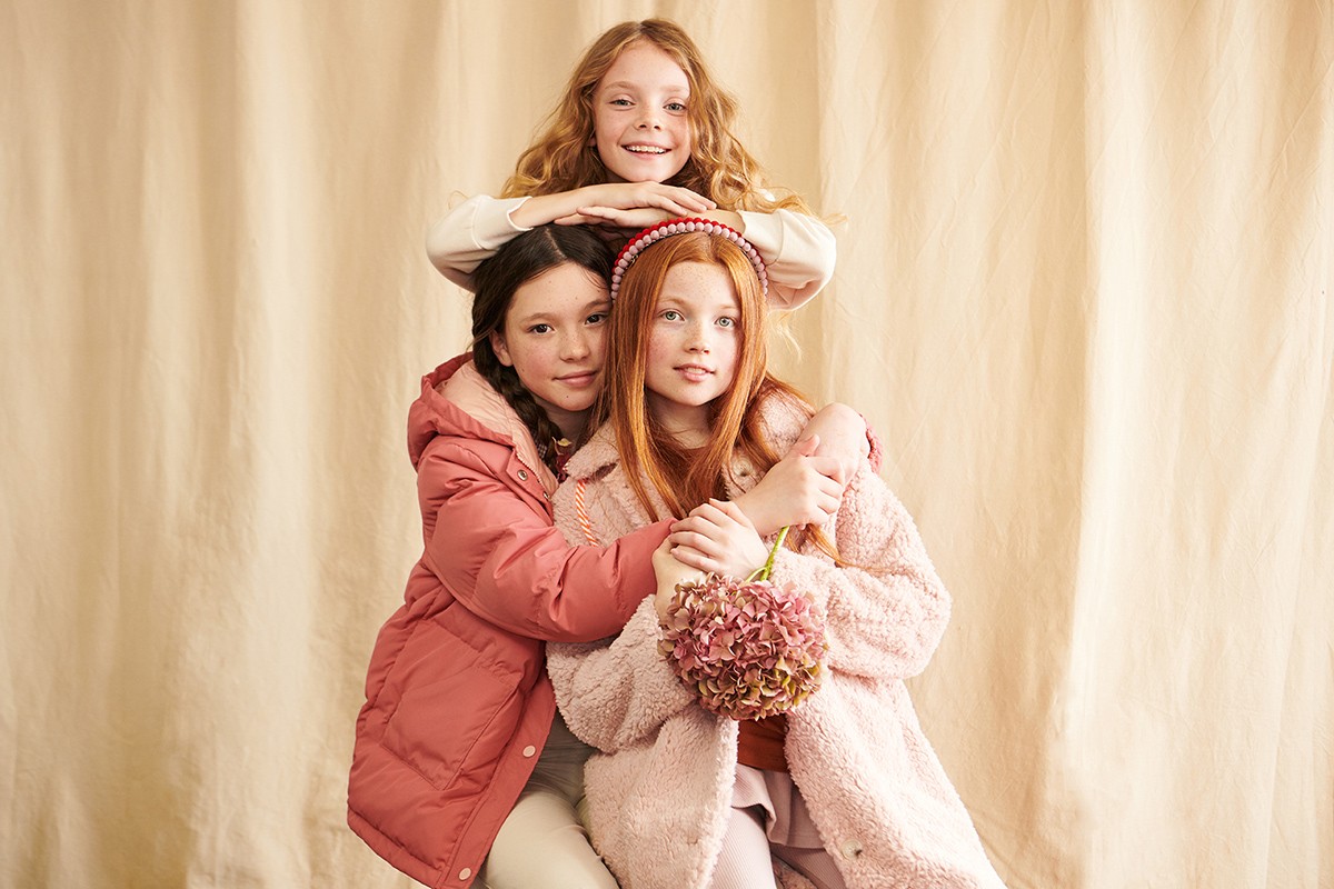 Three young girls in layered winter coats pose against a tan backdrop, displaying cozy fashion and sibling camaraderie indoors.
