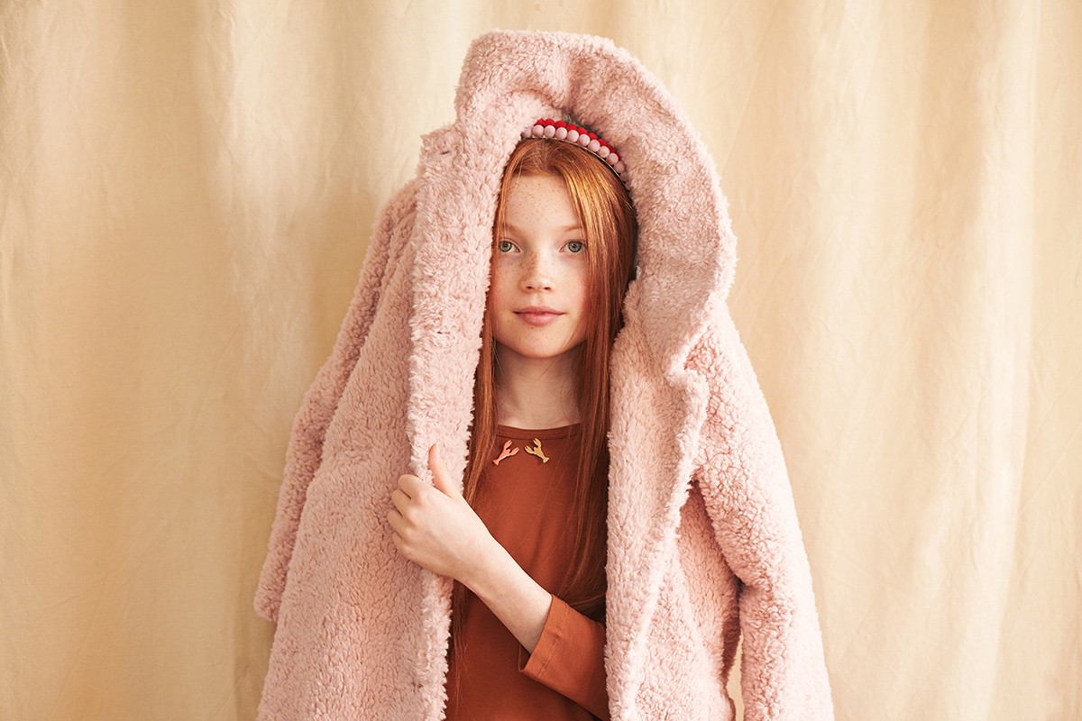 Red-haired child poses in a pink, fuzzy coat against a light tan backdrop, looking directly at the camera with a neutral expression.