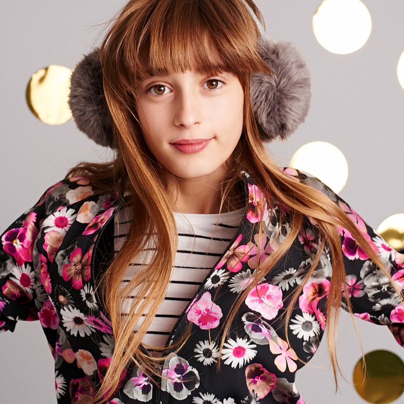 Portrait of a cheerful young girl wearing grey earmuffs and a black floral jacket, set against a grey background with gold bokeh circles.