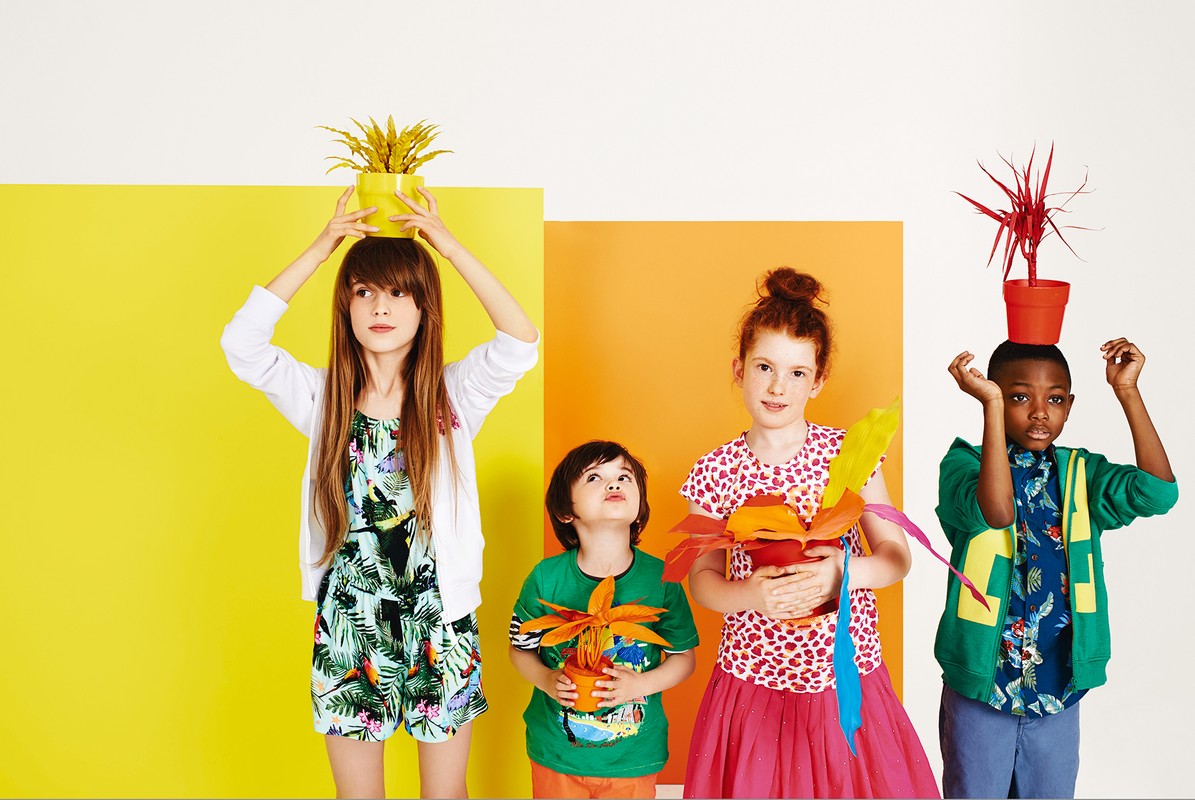 Four young children pose with colorful painted plants in pots against blocks of yellow and orange for a vibrant, playful fashion shot.