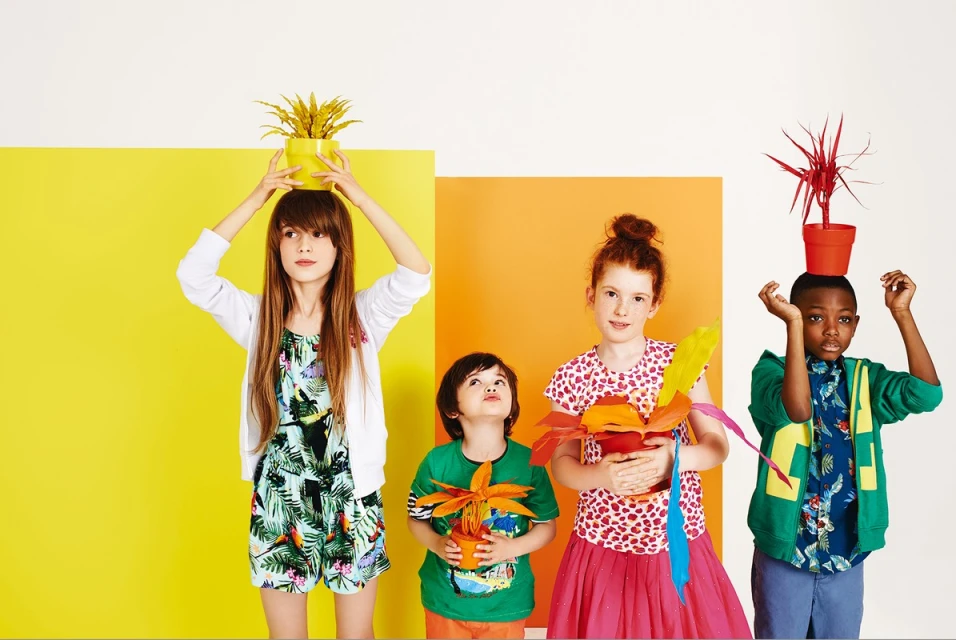 Four young children pose with colorful painted plants in pots against blocks of yellow and orange for a vibrant, playful fashion shot.