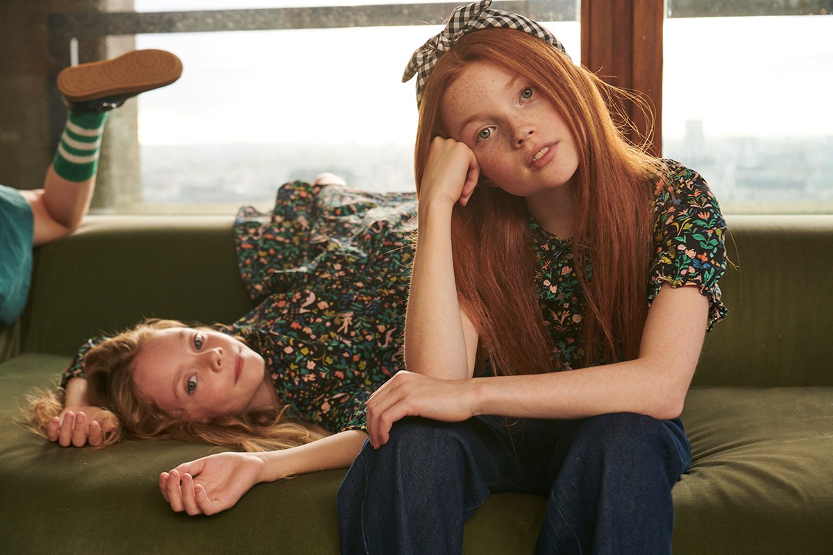 Two young girls pose on an olive-green couch in a high-rise apartment setting. One girl sits upright, while another reclines in the background.