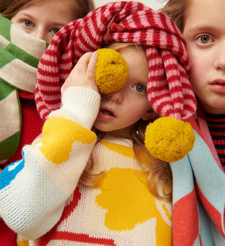Three children in bright, colorful knitwear pose indoors. One holds a yellow pompom to her eye, wearing a striped hat with pompoms.