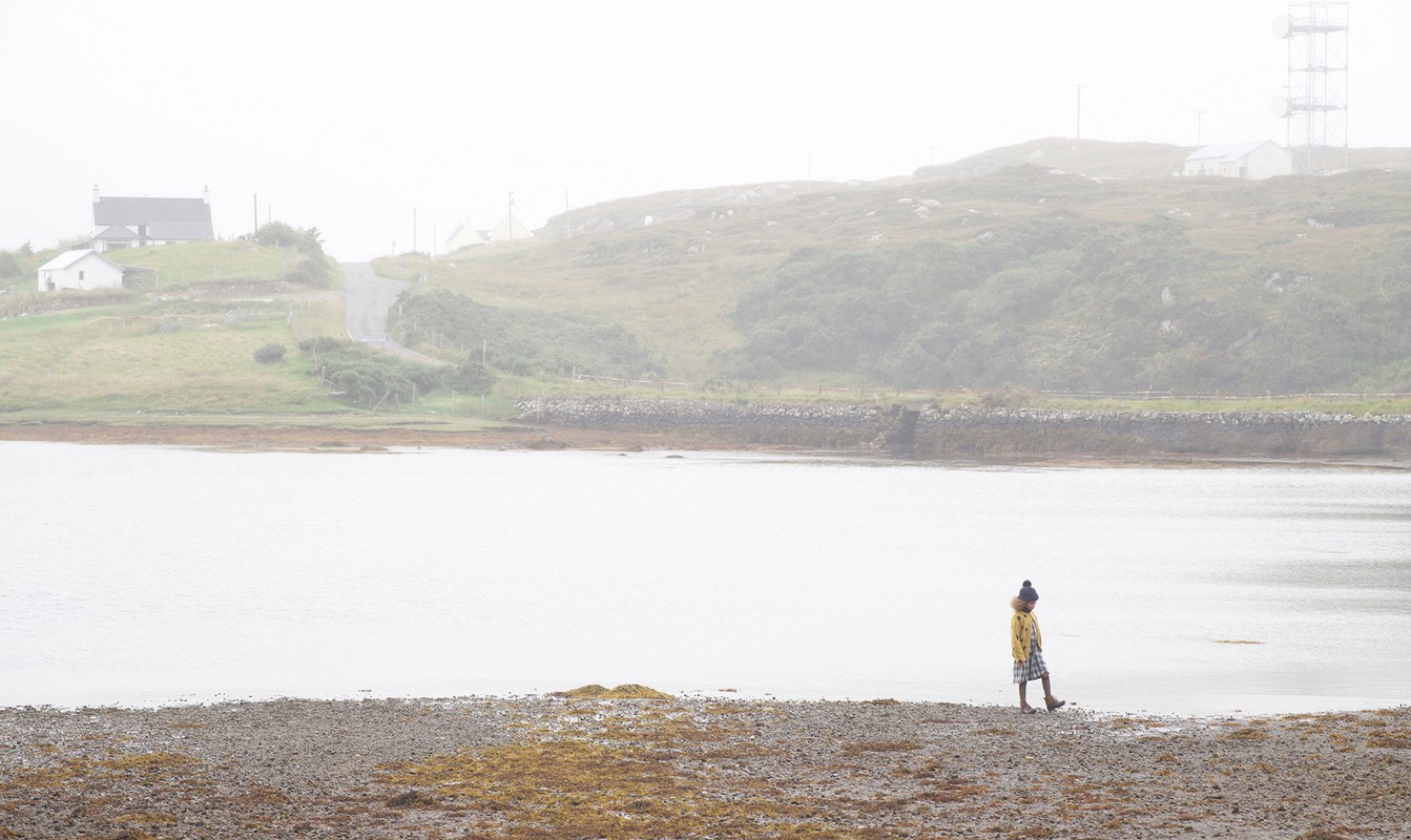 Child in yellow raincoat walking on a rocky beach in a misty rural landscape, with water, distant green hills and houses.