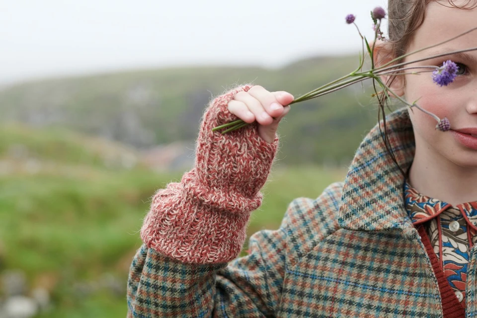 Young girl poses outdoors holding purple flowers, wearing a rustic, colorful wool coat and fingerless gloves in a misty environment.