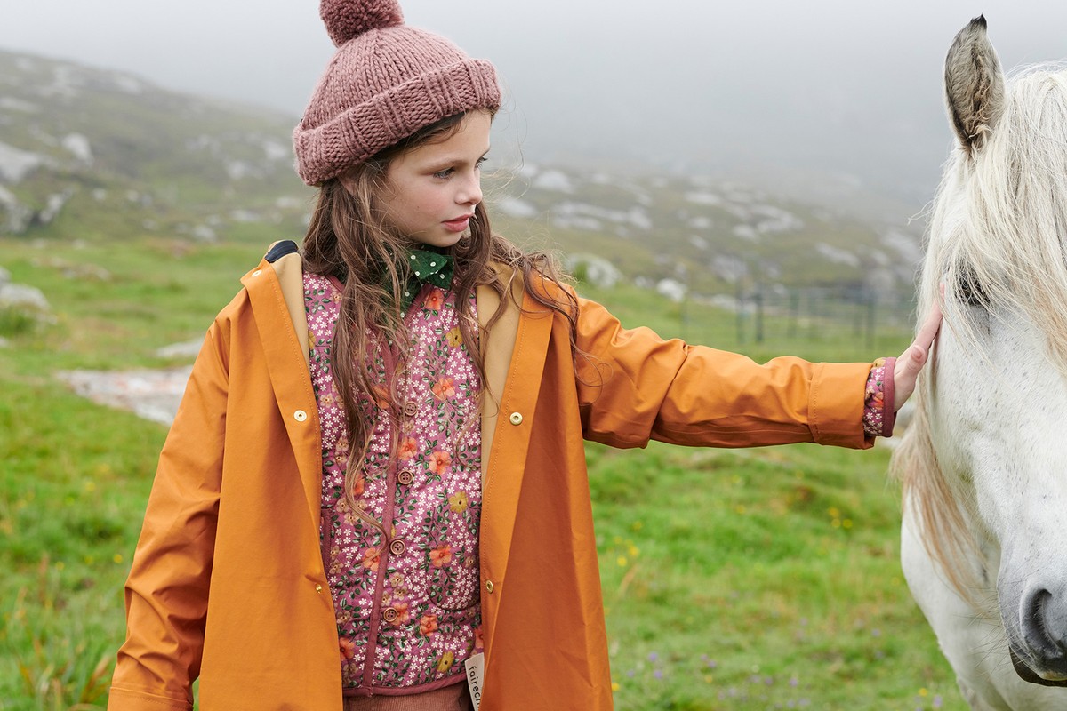 Young girl in orange raincoat interacts with a white horse in a lush, green field on a misty day, creating a serene outdoor scene.