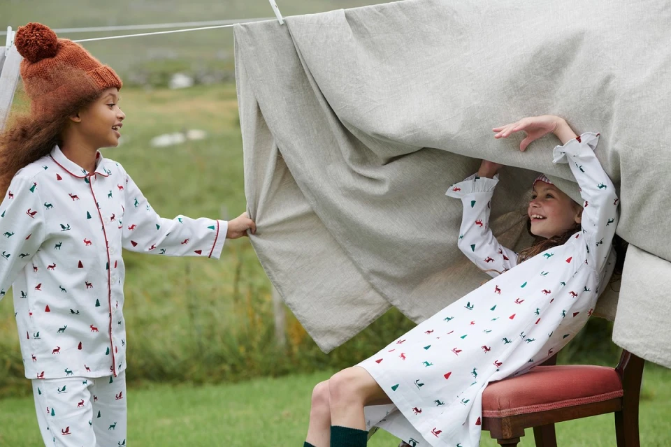 Two young girls play under a beige linen sheet hanging on a clothesline in a green field wearing matching Christmas-themed pajamas.