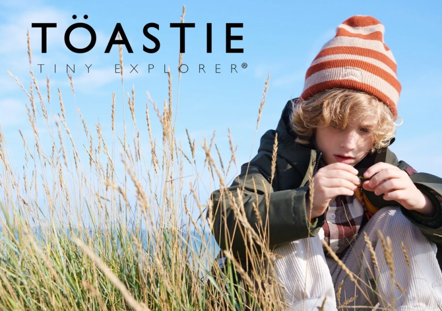 Child wearing a striped beanie and green jacket sits among tall grass under a blue sky in an outdoor kids fashion shot.