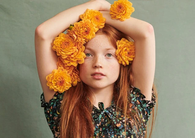 Red-haired young woman with freckles posing with bright yellow dahlia flowers tucked into her hair by photographer Joanna Paterson.