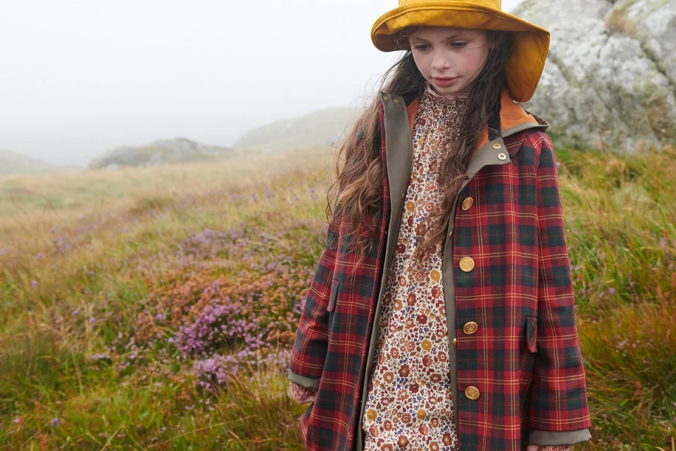 A young girl with long brown hair wears a plaid coat and yellow hat while standing in a grassy field with a misty mountain backdrop.
