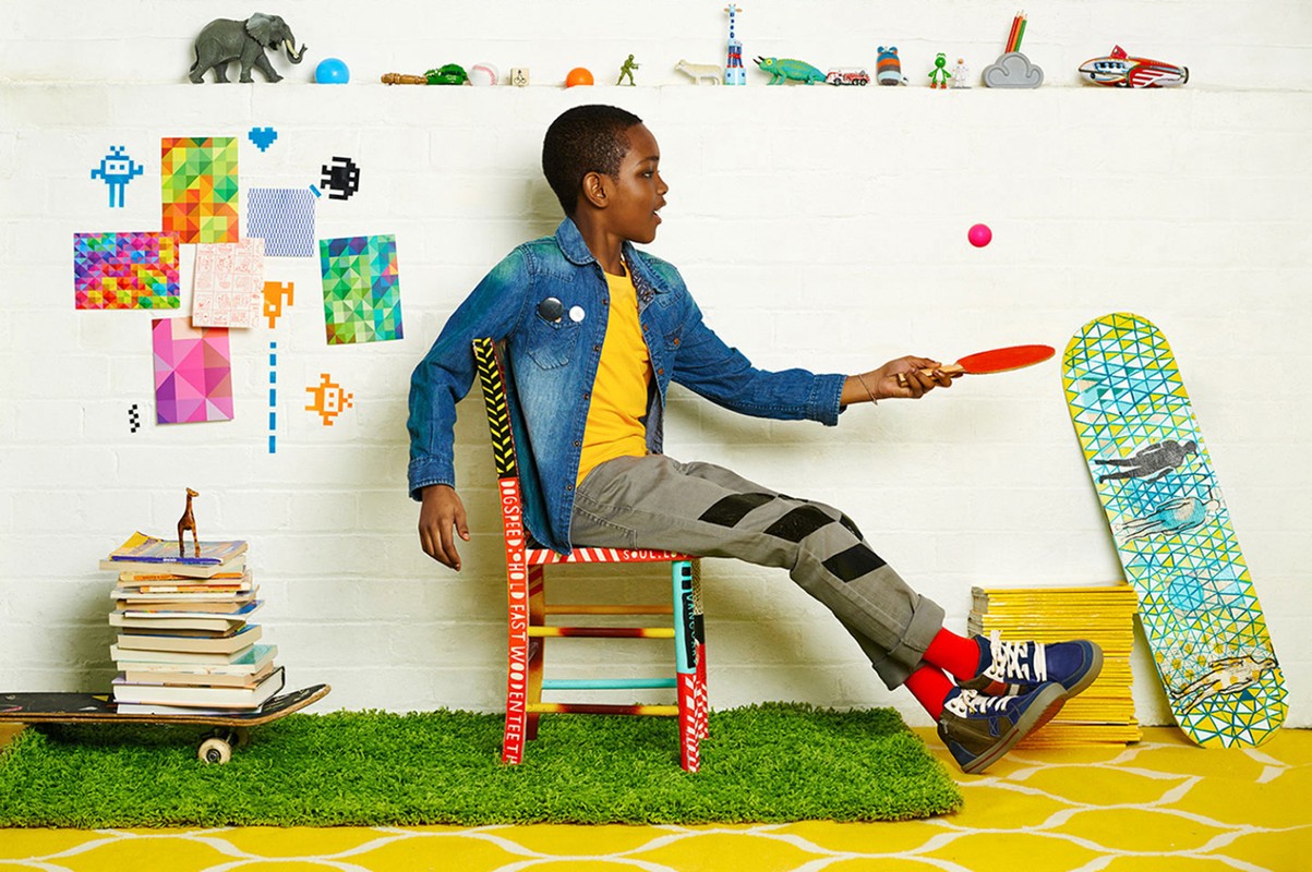 A young boy plays ping pong while sitting on a colorful chair in an indoor setting filled with toys, books, and playful decor.
